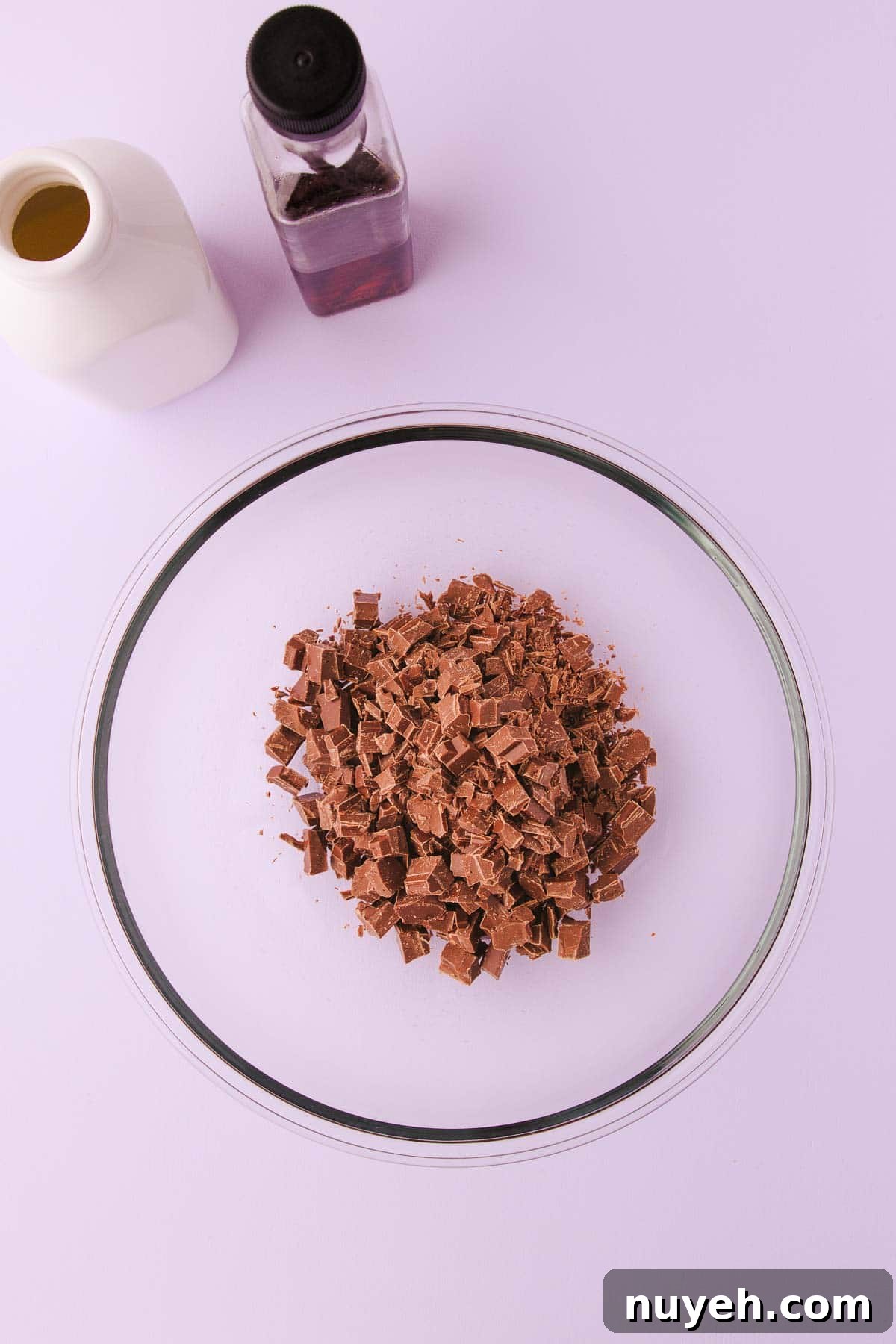 Chopped milk chocolate in a large glass bowl on a pale purple background.