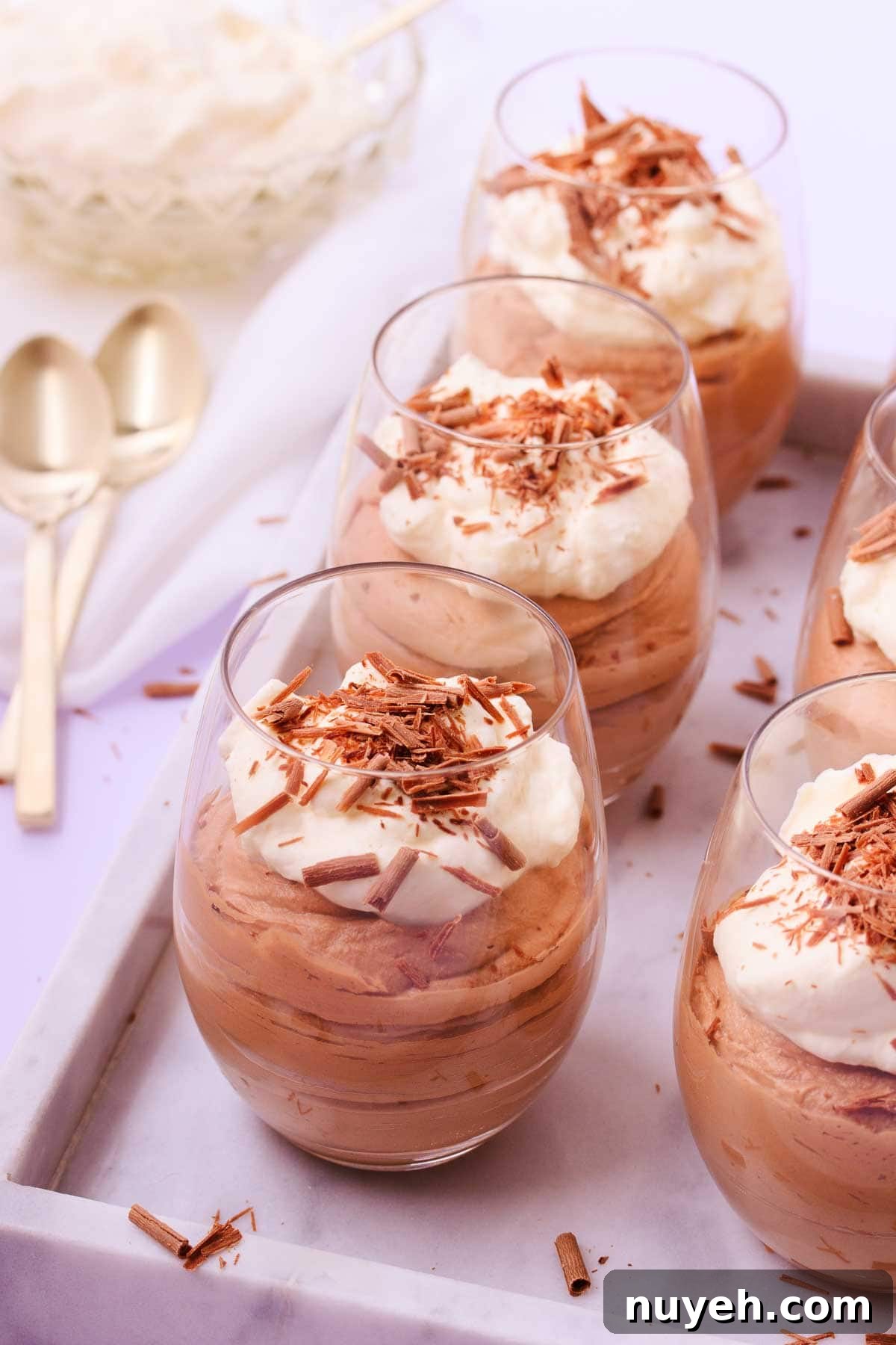 Six stemless wine glasses filled with milk chocolate mousse and topped with whipped cream and chocolate shavings, on an offset marble tray, with gold teaspoons.