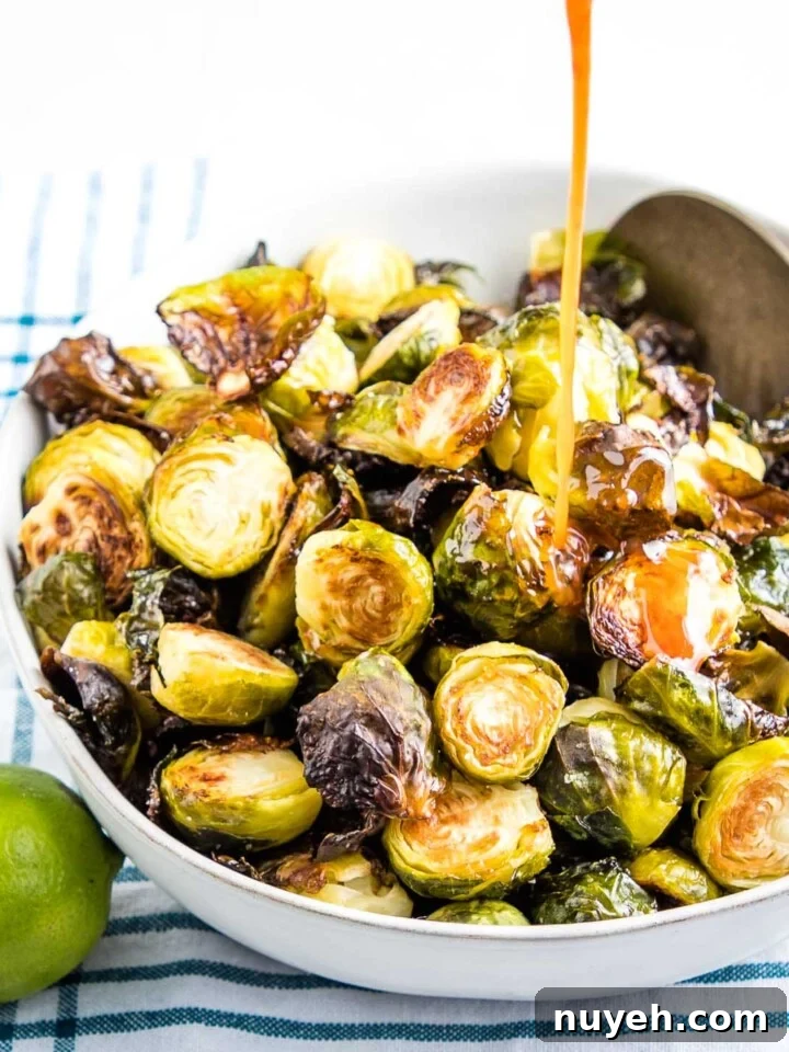 A stream of sweet and spicy sauce being poured onto a white bowl filled with crispy oven roasted brussels sprouts. There's a lime in the foreground and a teal striped towel underneath the bowl.