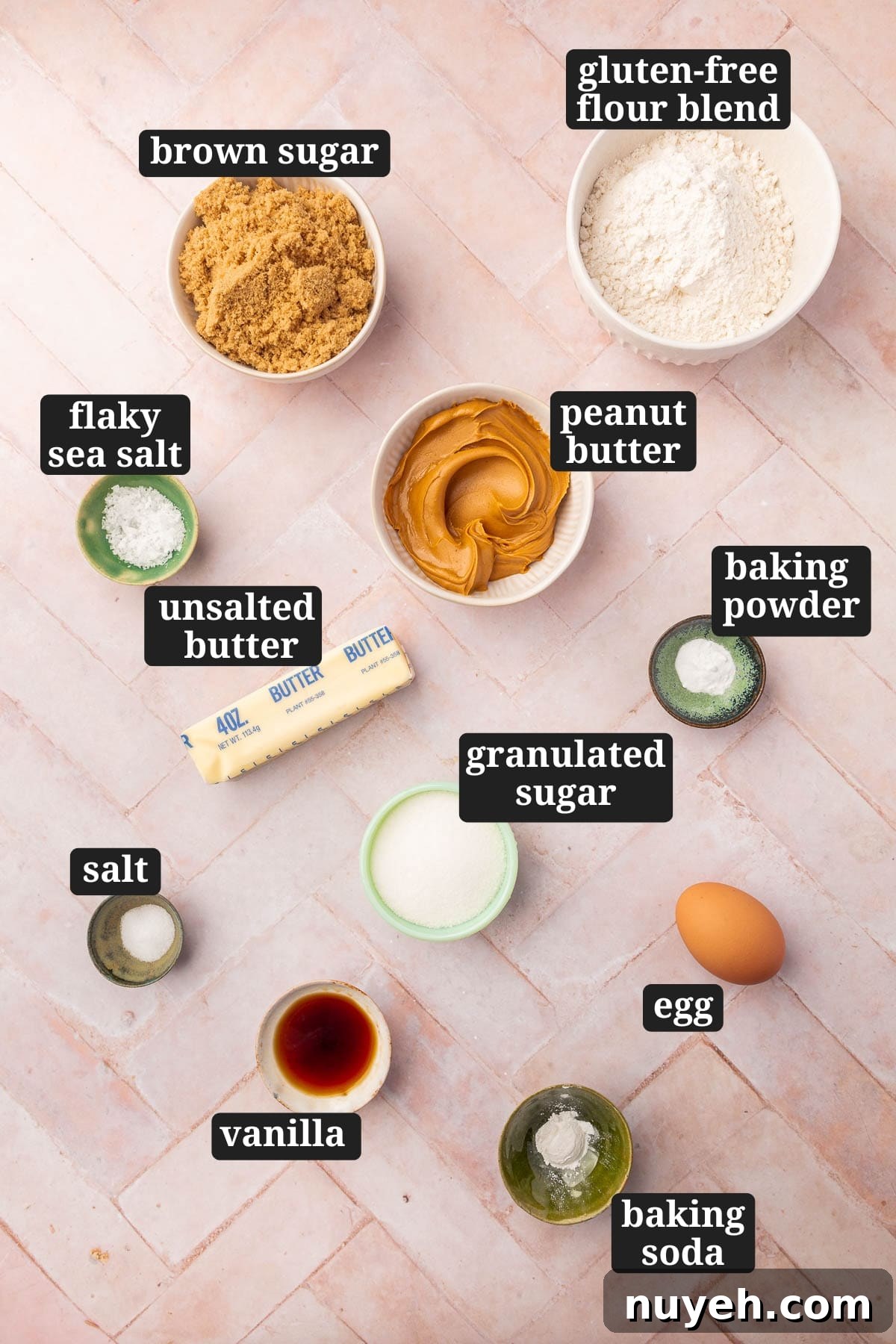 Ingredients in small bowls to make gluten-free peanut butter cookies, including brown sugar, gluten-free flour, flaky sea salt, peanut butter, butter, baking soda, baking powder, granulated sugar, vanilla, egg, and salt with text overlays over each ingredient.