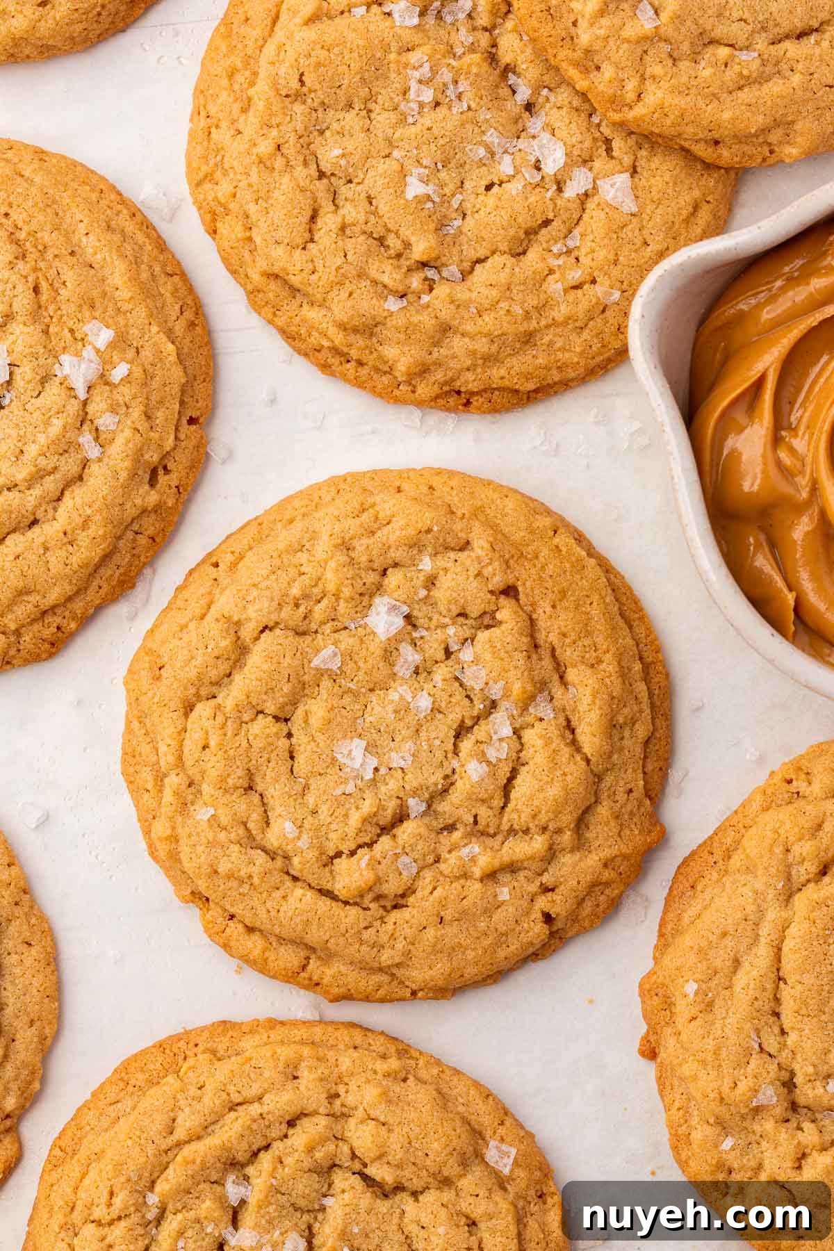 GF peanut butter cookies with flaky sea salt on a piece of parchment paper with a bowl of peanut butter peaking in.