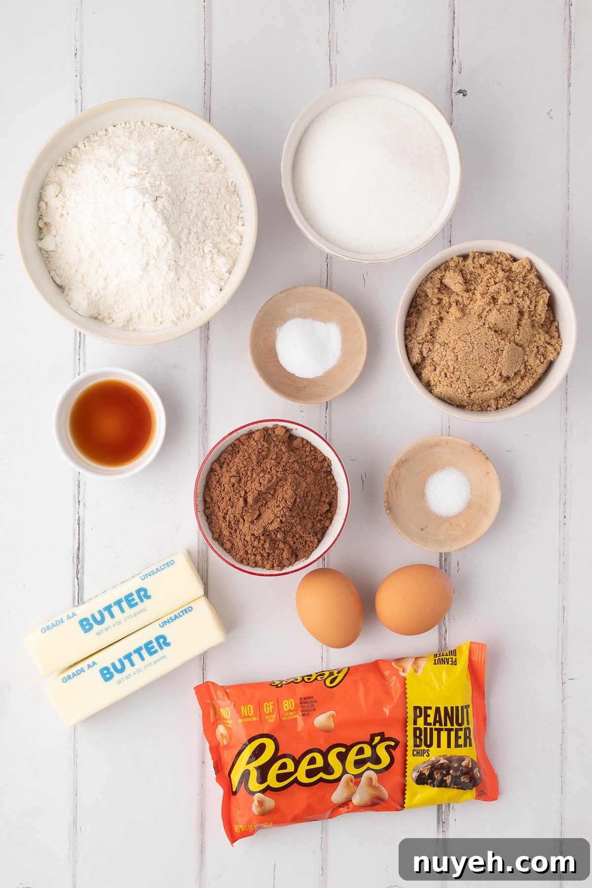 Overhead view of ingredients for chocolate peanut butter chip cookies, including unsweetened cocoa powder, flour, Reese's peanut butter chips, butter, and eggs on a white counter.