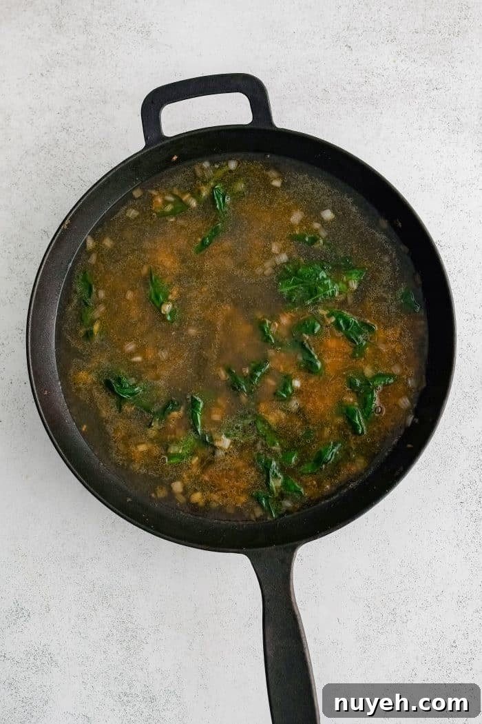 Large cast iron skillet filled with golden softened onions, garlic, and wilted fresh spinach in chicken broth.