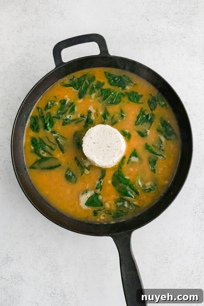 One wheel of Garlic and Herb Boursin cheese added to the center of a large cast iron skillet filled with golden softened onions, garlic, and wilted fresh spinach in chicken broth.