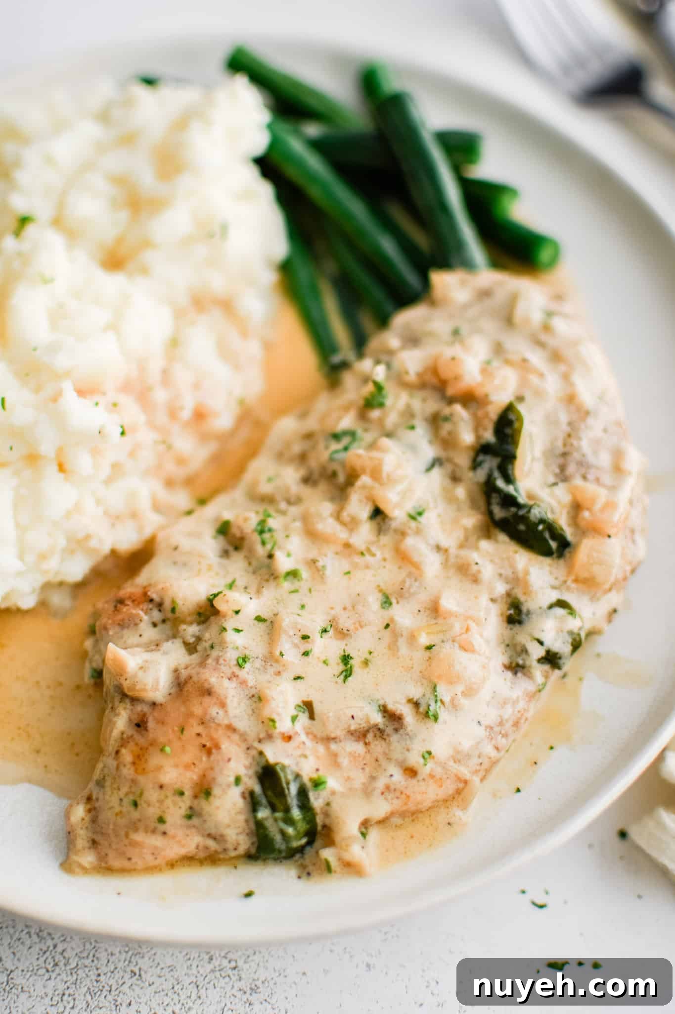 White serving plate filled with one piece of cooked chicken, a scoop of mashed potatoes, and pan-fried green beans all smothered in creamy boursin cheese sauce.