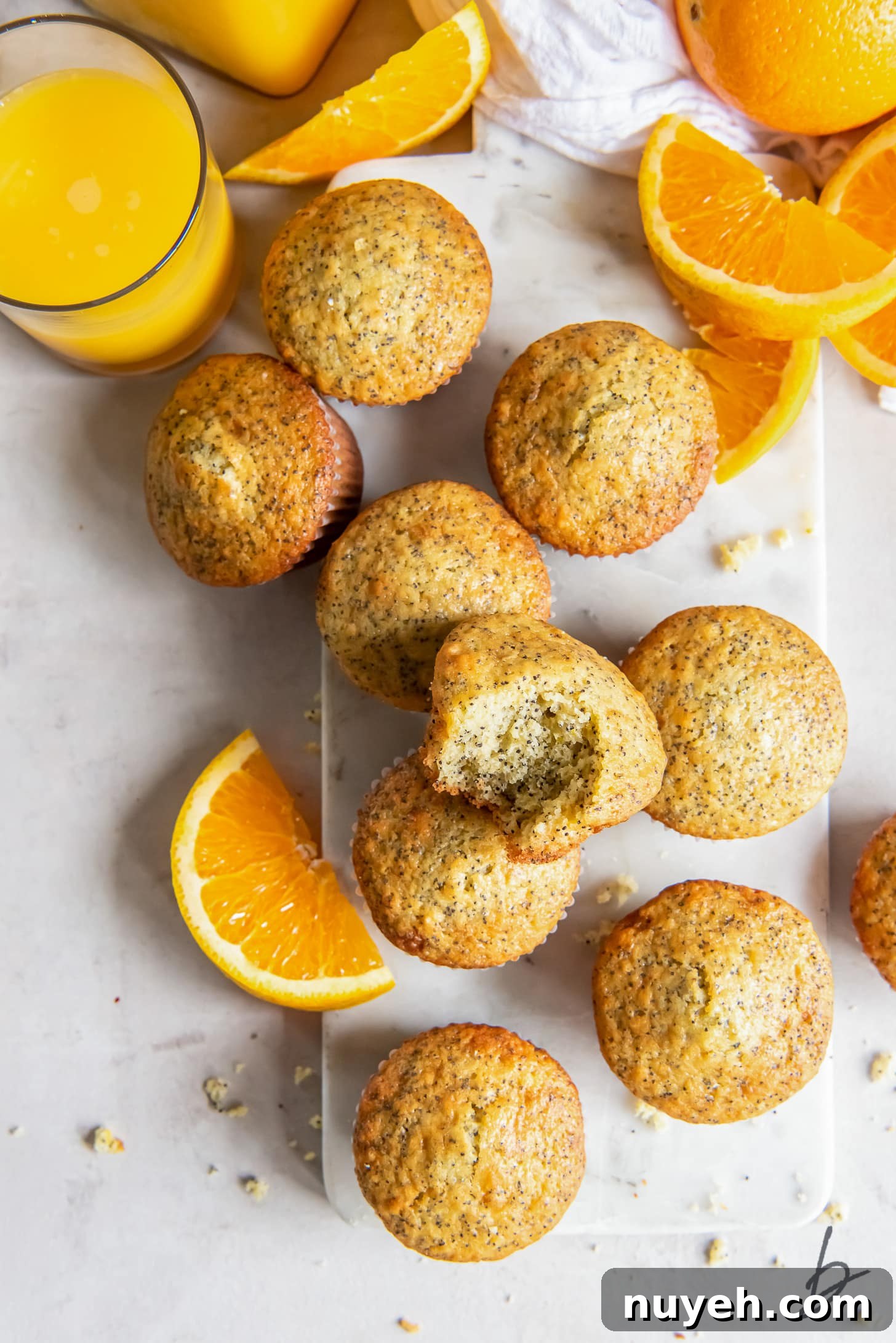 Zesty Orange Poppy Seed Muffins Recipe 3 orange poppy seed muffins with one muffin with a bite on a marble slab next to orange slices