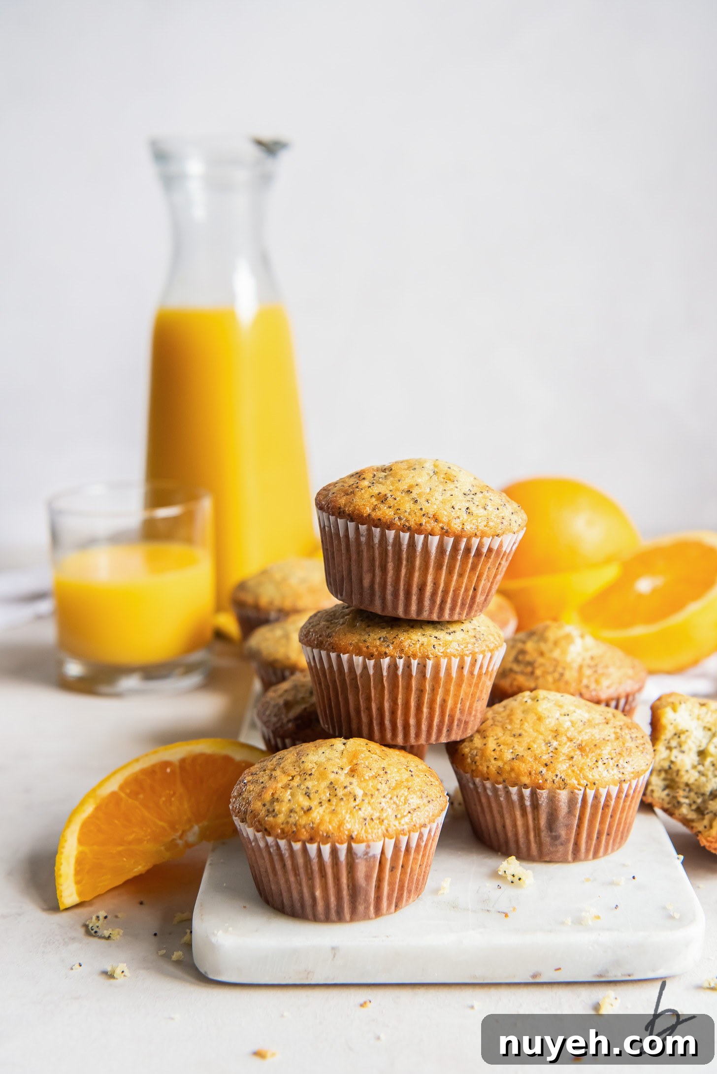 Zesty Orange Poppy Seed Muffins Recipe 6 orange poppy seed muffins stacked on top of each other in front of pitcher of orange juice and fresh oranges