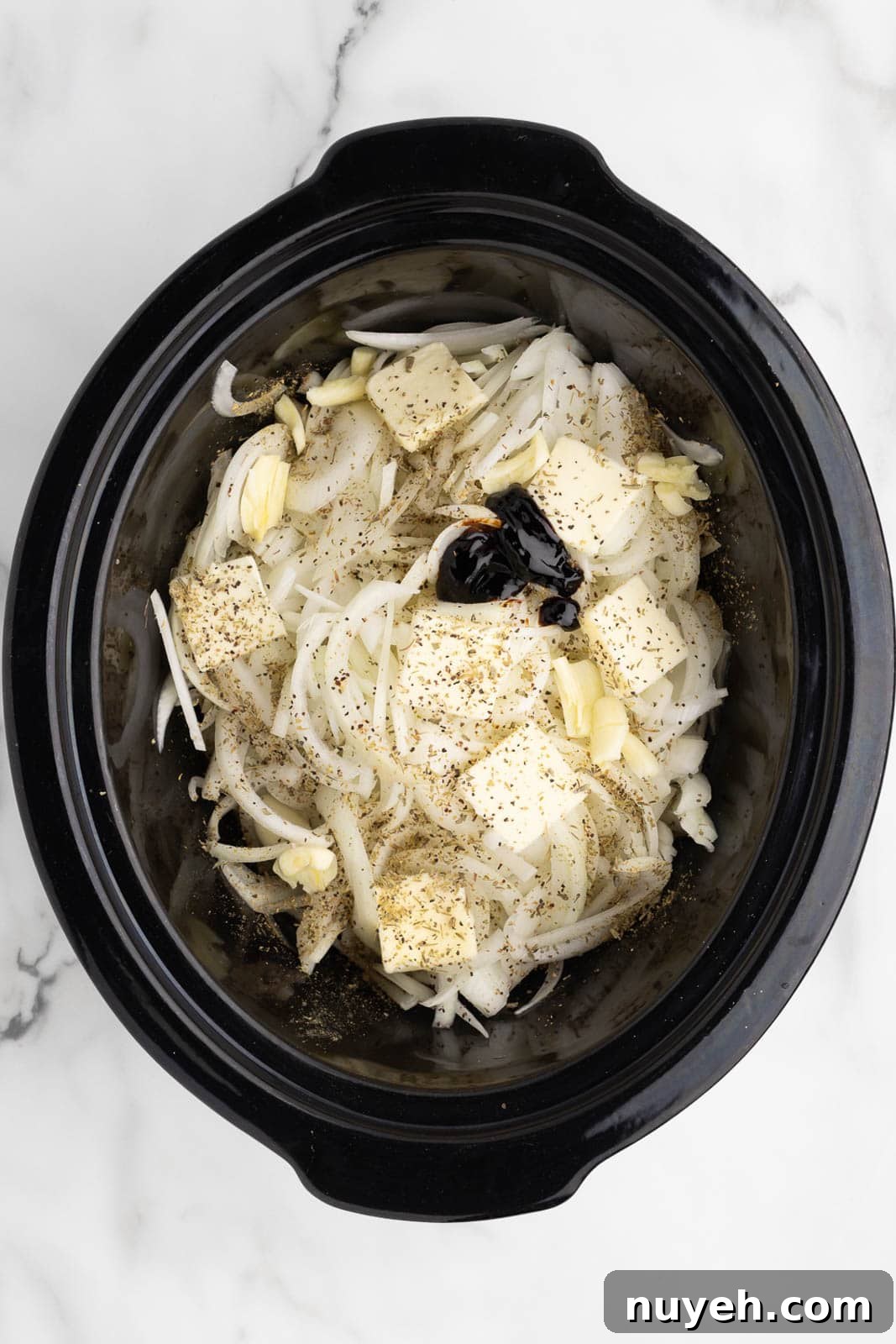black slow cooker of sliced onions covered with seasonings with butter and balsamic glaze