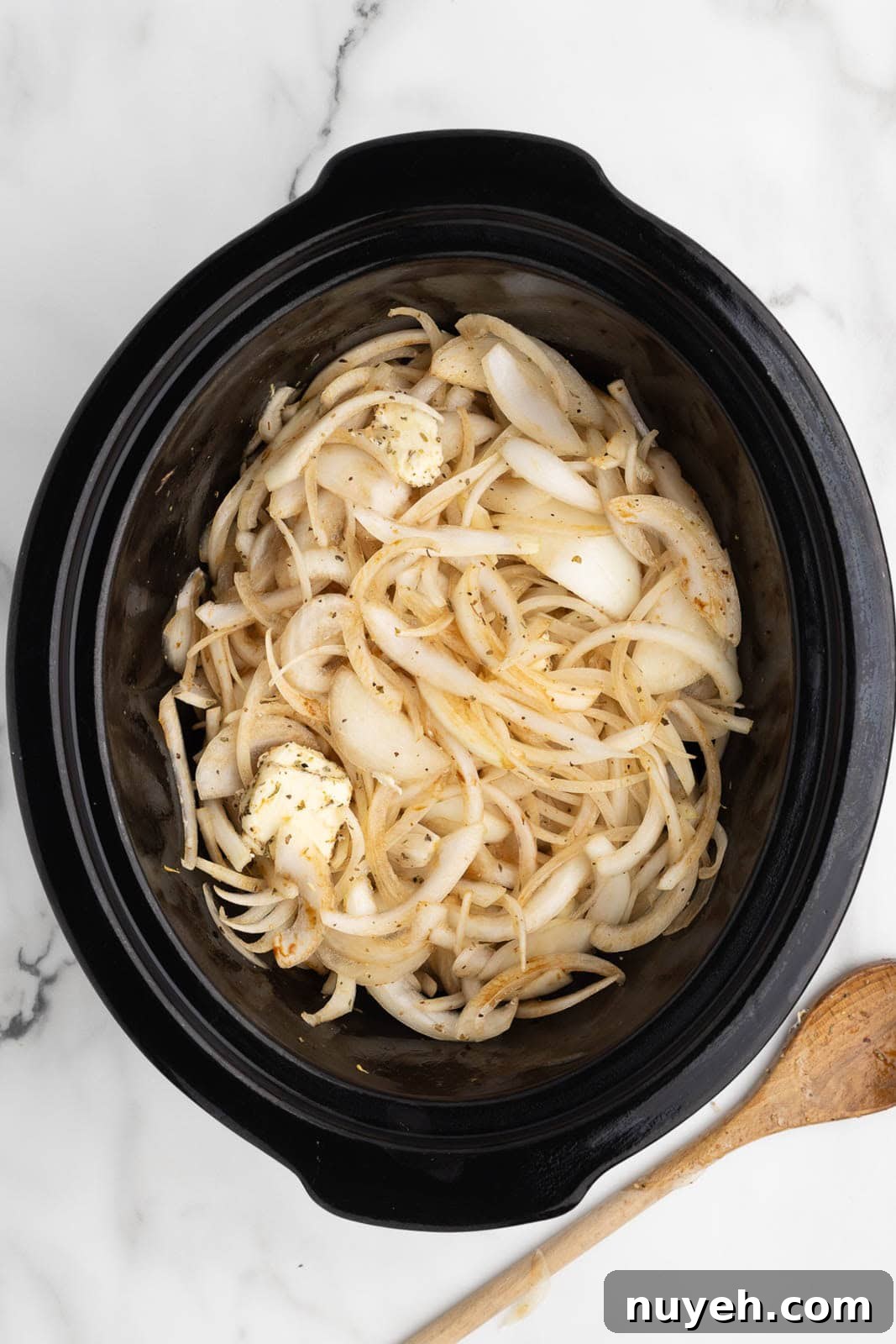 black slow cooker of sliced onions covered with seasonings with butter and balsamic glaze that have been stirred