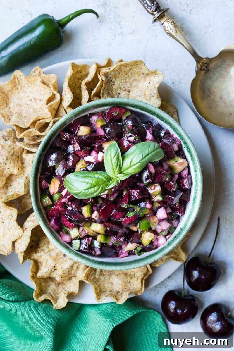 bowl of cherry salsa next to tortilla chips with green napkin, 3 cherries, jalapeno pepper, and serving spoon.